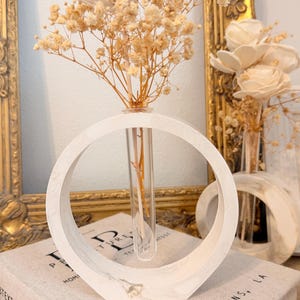 May include: A circular white marble-patterned stand with a central clear glass test tube vase. The vase holds dried beige baby's breath flowers and a single white wooden flower. A second, smaller matching circular stand is visible in the background.