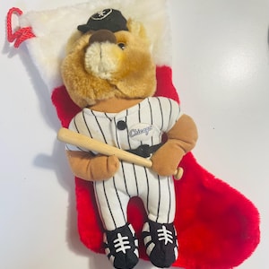 May include: A plush brown bear wearing a white and black striped baseball uniform with a wooden bat. The bear is wearing a black baseball cap with a white star on it. The bear is in a red and white stocking with the word "Chicago" on the uniform.