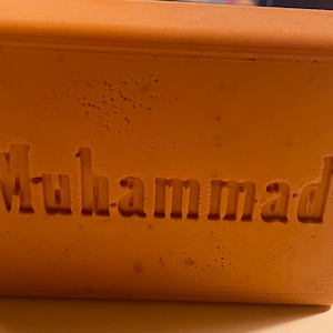 May include: A rectangular, orange-brown soap bar with the word "Muhammad" embossed on the front.