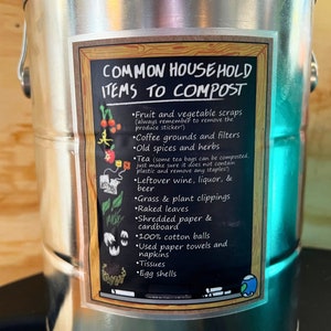 May include: A white sticker on a silver metal container lists common household items to compost. The sticker has a chalkboard background with a wooden frame and a green globe icon. The list includes fruit and vegetable scraps, coffee grounds and filters, old spices and herbs, tea bags, leftover wine, liquor, and beer, grass and plant clippings, raked leaves, shredded paper and cardboard, 100% cotton balls, used paper towels and napkins, tissues, and egg shells.