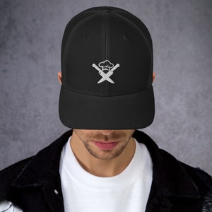 May include: Black trucker hat with a white embroidered chef's hat and crossed knives logo. The hat has a curved brim and a mesh back. A stylish accessory for culinary enthusiasts.
