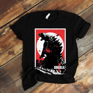 Op de afbeelding: Zwart T-shirt met een grafische print van Godzilla in een rood en wit posterontwerp. De poster heeft onderaan de tekst "Godzilla".