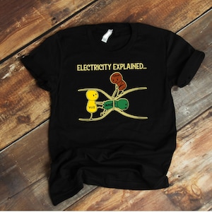 Pode incluir: Camiseta preta com um gráfico de uma ilustração de desenho animado que explica a eletricidade. A ilustração mostra uma pessoa rotulada como "Ohm" segurando duas pessoas rotuladas como "Volt" e "Amp" em um fio.