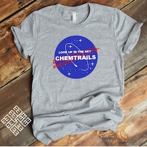 Puede incluir: Una camiseta gris con un gráfico circular azul que dice "Look up in the sky Chemtrails". El gráfico está en el estilo de un logotipo de la NASA.