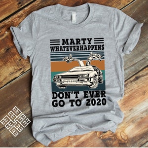 以下が含まれることがあります： 映画「バック・トゥ・ザ・フューチャー」のデロリアンタイムマシンをヴィンテージスタイルのグラフィックで描いたグレーのTシャツ。グラフィックには「Marty Whatever Happens Don't Ever Go To 2020」というテキストが含まれています。