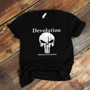 Può includere: Maglietta nera con una grafica bianca di un teschio con un design di bandiera americana. Il testo "Devolution" e "Patriots are in control" è stampato sulla maglietta.