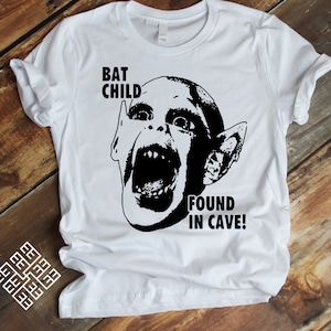 May include: White t-shirt with a black and white graphic of a creature with large ears and the text "BAT CHILD FOUND IN CAVE!"