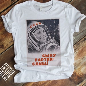 May include: White t-shirt featuring a vintage Soviet space propaganda poster. The poster depicts a smiling cosmonaut in a spacesuit against a starry background, with Cyrillic text. The text reads "CCCP" and "Сыну партии - слава!"