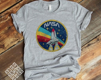 Peut inclure: Un t-shirt gris avec un logo vintage de la NASA représentant une navette spatiale décollant sur un fond bleu, rouge, jaune et turquoise avec des étoiles.