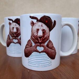 Può includere: Due tazze in ceramica bianca con un orso marrone che indossa una camicia a righe blu e bianche e un cappello rosso con una stella. L'orso sta formando un cuore con le sue zampe.