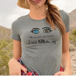 Op de afbeelding: Grijs T-shirt met een afbeelding van twee ogen, één blauw en één bruin. De tekst luidt "Laat me je vraag beantwoorden: Ik ben zo geboren."