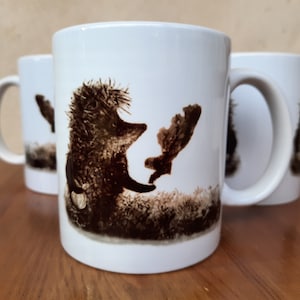 Puede incluir: Taza de cerámica blanca con una ilustración marrón de un erizo sosteniendo una hoja. El erizo está mirando hacia la izquierda y la hoja hacia la derecha. El fondo es una ilustración marrón y blanca de hierba y hojas.