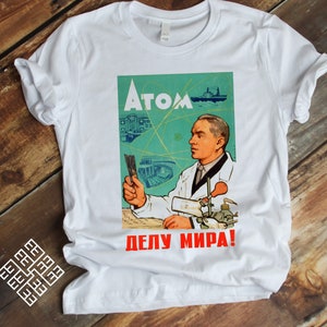 May include: White t-shirt with a vintage-style graphic of a scientist working in a lab. The graphic features the text "Atom Делу Мира!" which translates to "Atom For the Cause of Peace!"