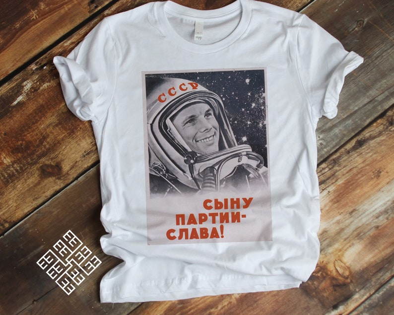 May include: White t-shirt featuring a vintage Soviet space propaganda poster. The poster depicts a smiling cosmonaut in a spacesuit against a starry background, with Cyrillic text. The text reads "CCCP" and "Сыну партии - слава!"