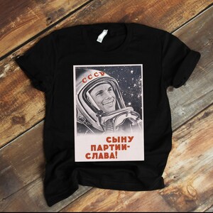 May include: Black t-shirt featuring a vintage Soviet space propaganda poster. The poster shows a smiling cosmonaut in a space helmet against a starry background, with the word "CCCP" and Cyrillic text. A classic design.