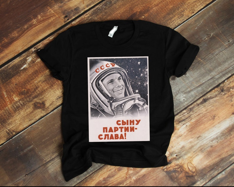 May include: Black t-shirt featuring a vintage Soviet space propaganda poster. The poster shows a smiling cosmonaut in a space helmet against a starry background, with the word "CCCP" and Cyrillic text. A classic design.