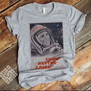 May include: A gray t-shirt featuring a vintage image of a smiling cosmonaut in a space helmet. The image includes the word "CCCP" in red and Russian text. The shirt is displayed on a wooden surface.