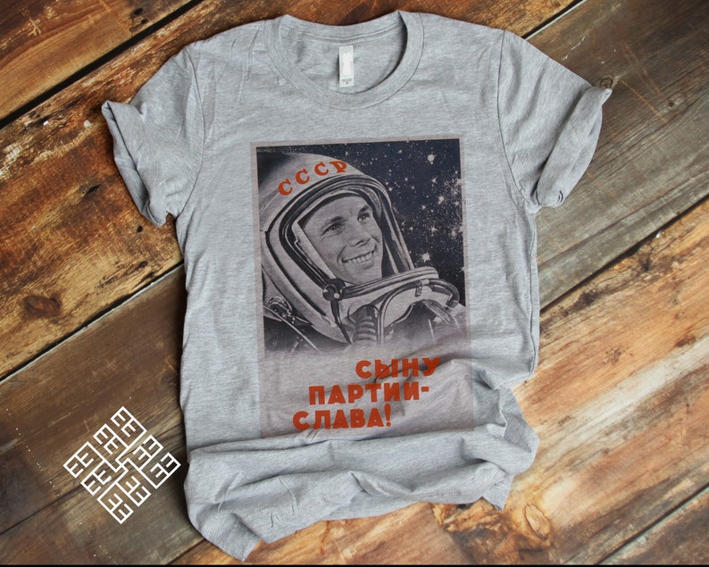 May include: A gray t-shirt featuring a vintage image of a smiling cosmonaut in a space helmet. The image includes the word "CCCP" in red and Russian text. The shirt is displayed on a wooden surface.