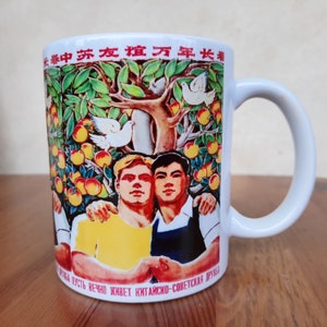 Peut inclure: Une tasse en céramique blanche avec une image colorée de deux hommes se serrant la main devant un arbre avec des oiseaux et des oranges. Le texte sur la tasse est "*春中苏友谊万年长者*" et "МИНЬ ПУСТЬ ВЕЧНО ЖИВЕТ КИТАЙСКО-СОВЕТСКАЯ ДРУЖБА".