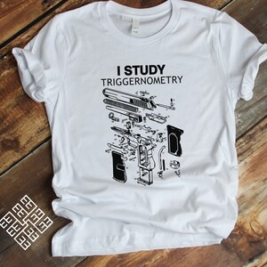 May include: White t-shirt featuring a black and white illustration of a disassembled handgun and the text "I study triggernometry".