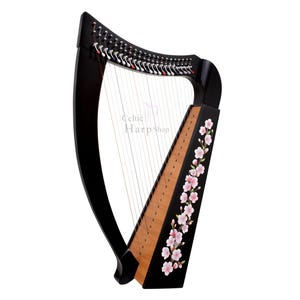 May include: A black Celtic harp with strings, featuring a wooden soundboard and pink floral accents. The harp has a curved pillar and an elegant design. The text "Celtic Harp Shop" is visible.