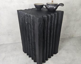 May include: A black, geometric side table with a textured surface. A small, dark tea set, including a teapot and a bowl, sits on top. The table is made of wood and has a unique, modern design.