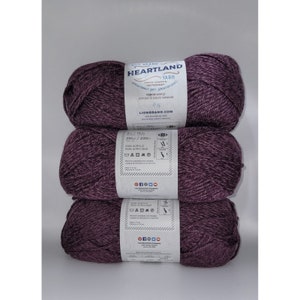 May include: Three skeins of purple Heartland yarn, a Lion Brand yarn. The yarn is 100% acrylic and comes in a 251-yard skein. The label includes care instructions and social media icons.