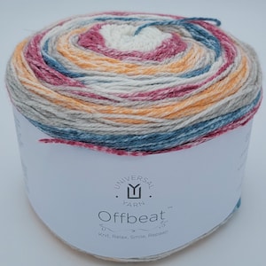 Puede incluir: Una madeja de hilo en un patrón multicolor de naranja, rosa, azul y blanco. El hilo está etiquetado como "Universal Yarn Offbeat" y el lema "Knit, Relax, Smile, Repeat!"