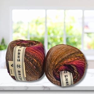 Pode incluir: Dois novelos de lã redondos com cores variadas, apresentando uma mistura de fibras castanhas, cor de laranja, roxas e pretas. Cada novelo está envolvido por uma etiqueta de papel creme impressa com texto japonês preto e um logótipo circular com a letra N.