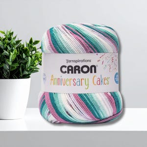 Pode incluir: Um novelo de fio Caron Anniversary Cakes em tons de verde-azulado, rosa, branco e lavanda. O fio é rotulado com o nome da marca e do produto. Uma pequena planta em vaso está à esquerda do fio em uma superfície branca.