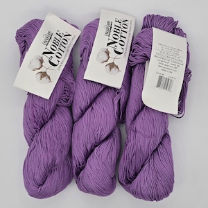 Pode incluir: Três novelos de fio Noble Cotton roxo com etiquetas brancas anexadas. As etiquetas exibem o nome da marca, o tipo de algodão e uma ilustração de planta de algodão. Cada novelo tem 200 g.
