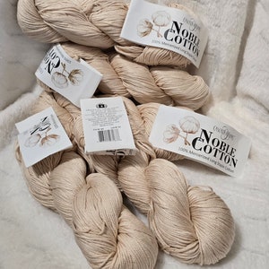 Pode incluir: Vários novelos de fio Noble Cotton da Cascade Yarns, 100% algodão mercerizado de fibra longa. Cada novelo tem um rótulo com o nome da marca e uma ilustração de flor de algodão. O fio está sobre uma superfície branca e macia.