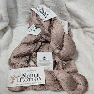 Pode incluir: Vários novelos de Cascade Yarns Noble Cotton em um tom rosa suave. Cada novelo tem uma etiqueta que diz "Noble Cotton" e "100% Mercerized Long Staple Cotton". O fio é leve, com 200 m por novelo de 100 g.