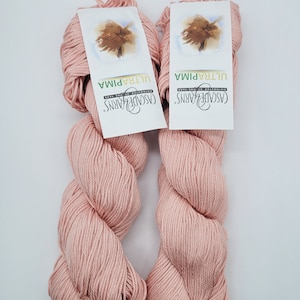 Pode incluir: Dois novelos de fio rosa Cascade Yarns Ultra Pima, etiquetados como "Ultra Pima" e "Cascade Yarns". O fio é de cor rosa suave e claro.