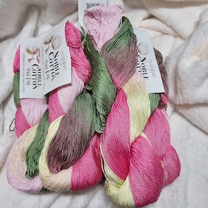 May include: A collection of hand-dyed Noble Cotton yarn skeins in shades of pink, green, yellow, and brown. The yarn is labeled with the text "CASCADE YARNS NOBLE COTTON HAND DYED 100% Mercerized Long Staple Cotton". The skeins are arranged on a white, textured surface.