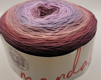 May include: A round ball of Lion Brand Mandala yarn in a gradient of colors, including shades of purple, pink, and burgundy. The yarn is wrapped in a paper label with the brand name and the word "mandala" in red script.