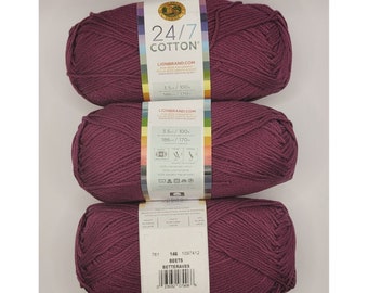 Könnte beinhalten: Drei Knäuel Lion Brand 24/7 Cotton Garn in der Farbe Rote Bete. Das Garn ist ein tiefes Burgunderrot. Das Etikett zeigt, dass das Garn aus 100% Baumwolle besteht und maschinenwaschbar ist.