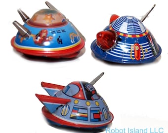 May include: Three vintage tin toy spaceships in blue, red, and silver. The toys have a retro design with details like fins, lights, and painted illustrations. The toys are on a white background.
