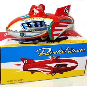 May include: A red and white wind-up toy rocket racer with a driver in the cockpit. The toy is on a blue and yellow box with the words "Rocket Racer" printed on it. The toy has a number "6" on the side and the words "Pocket Racer" on the top.