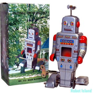 May include: A silver tin toy robot with red accents, a wind-up key on its side, and wheels at its base. The robot features a rectangular chest panel with colorful graphics and a small antenna on its head. It stands next to its original box.