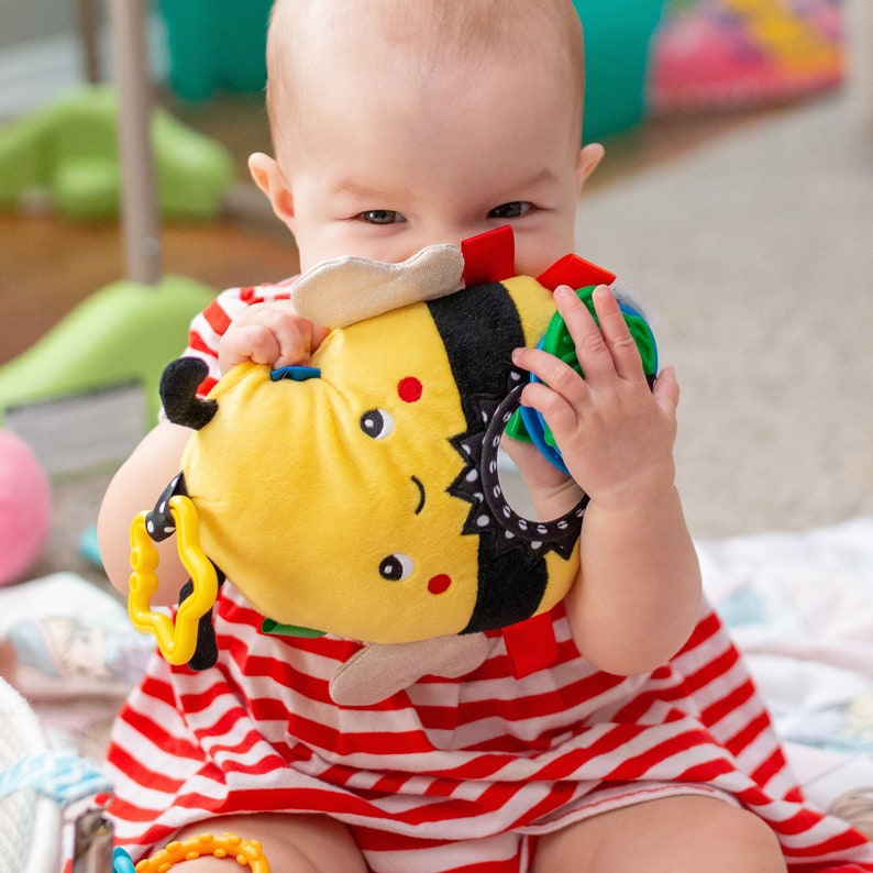 Op de afbeelding: Een baby in een rood-wit gestreepte jurk houdt een geel en zwart bij-vormig speelgoed vast met kleurrijke textuurdelen. Het speelgoed heeft een spiegel en verschillende texturen voor sensorisch spel.