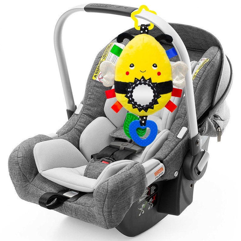 Op de afbeelding: Een grijze baby autostoel met een zwart en geel bij-vormig speelgoed dat aan de bovenkant hangt. Het speelgoed heeft een spiegel en een hartvormige bijtring.