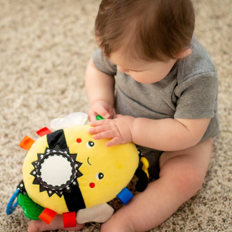Op de afbeelding: Een baby speelt met een geel en zwart bij-vormig pluche speelgoed met een spiegel en kleurrijke labels. Het speelgoed heeft een glimlachend gezicht en een zwart-wit gestreept patroon.
