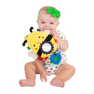 May include: A baby wearing a white and colorful onesie with a green bow in their hair is holding a yellow and black bee-shaped plush toy with a mirror and a heart-shaped teether.