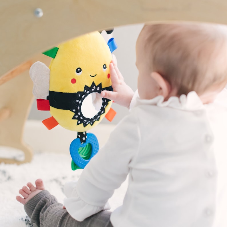 Op de afbeelding: Een geel en zwart bij-vormig babyspeelgoed met een spiegel en kleurrijke, textuurrijke ringen. Het speelgoed hangt aan een houten constructie en een baby reikt ernaar.
