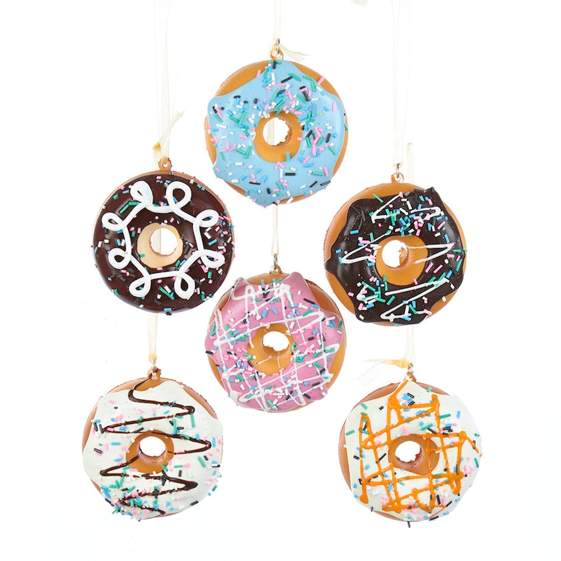 May include: Six donut shaped ornaments hanging from white strings. The donuts are decorated with different colored icings and sprinkles. The donuts are blue, brown, pink, white, and orange.