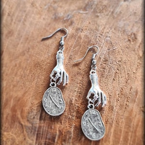 May include: A pair of silver earrings featuring a hand holding a clock face. The clock face is a silver oval with black Roman numeral markings.