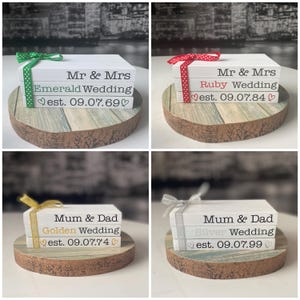 May include: Four white wooden blocks stacked on top of each other with a ribbon tied around the top block. The blocks are decorated with text that says "Mr & Mrs Emerald Wedding est. 09.07.69", "Mr & Mrs Ruby Wedding est. 09.07.84", "Mum & Dad Golden Wedding est. 09.07.74", and "Mum & Dad Silver Wedding est. 09.07.99". The blocks are sitting on a wooden base.