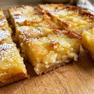 May include: Square-cut lemon bars with a golden-brown crust and a bright yellow, fruit-filled center, topped with a light dusting of white powdered sugar. The bars are arranged in a grid pattern on a wooden surface, showing a dense, baked texture.