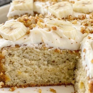 May include: A close-up of a banana cake with a layer of white icing and chopped pecans. The cake is cut in half, revealing the moist, yellow cake inside.